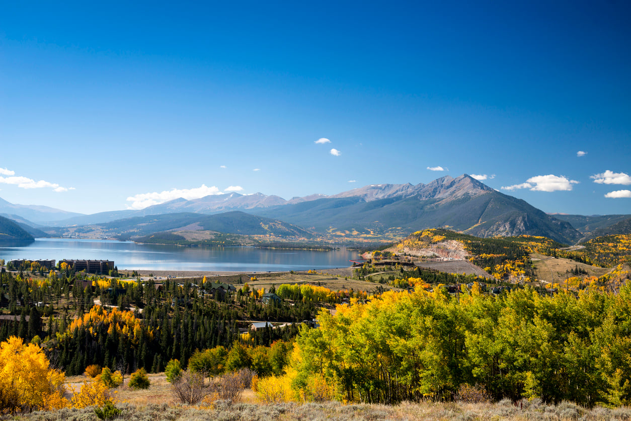 A scenic landscape reminiscent of the best fall towns in America, with a lake, mountains in the background, and vibrant yellow and green autumn trees under a clear blue sky.