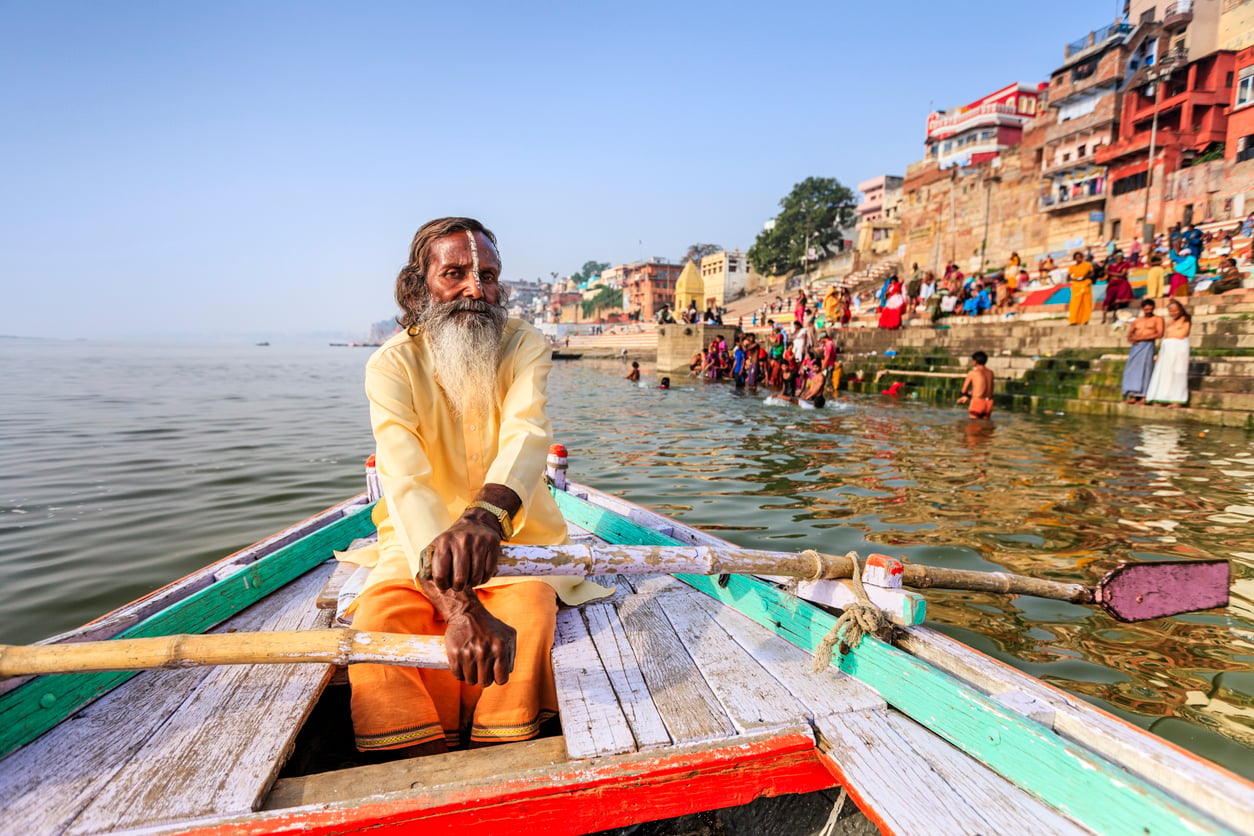An elderly man with a long beard rows a wooden boat on the Ganges River near colorful ghats in Varanasi, India, as people gather and bathe on the riverbank.