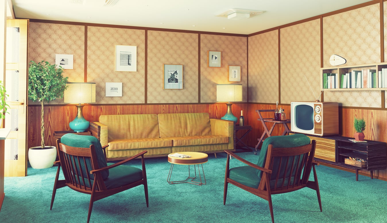 A retro living room with a yellow sofa, two wooden armchairs, a vintage TV, lamps, framed art on patterned walls, bookshelves, a potted plant, and teal carpet.