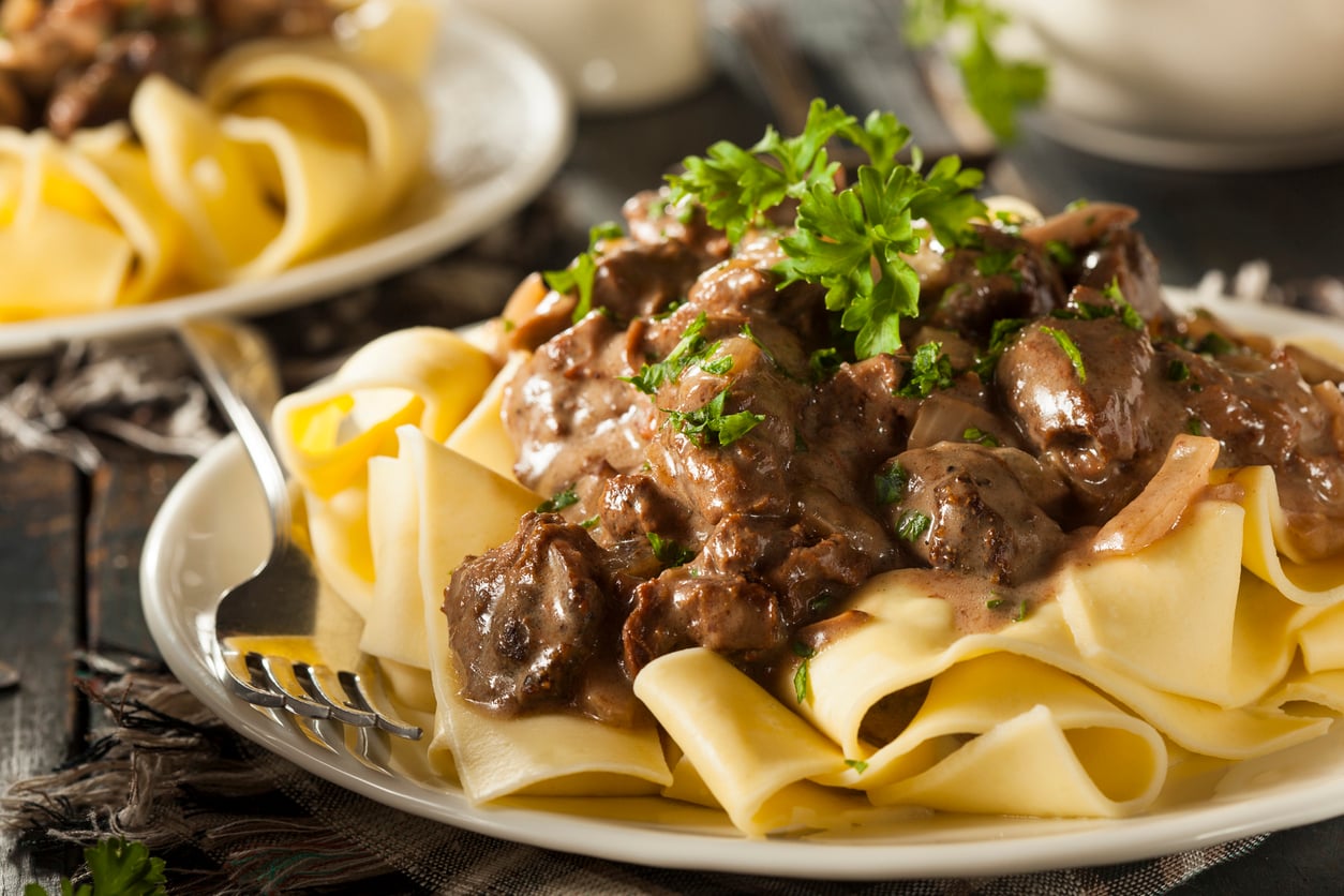 Wide egg noodles topped with creamy beef stroganoff and garnished with fresh parsley, served on a white plate with a fork beside the dish—just like the classic meals middle-class moms made for comforting family dinners.