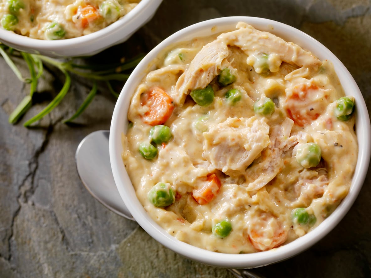 A bowl of creamy chicken pot pie filling with chunks of chicken, peas, and carrots, placed on a stone surface with a spoon beside it evokes the comforting meals middle-class moms made. Another partial bowl is visible in the background.