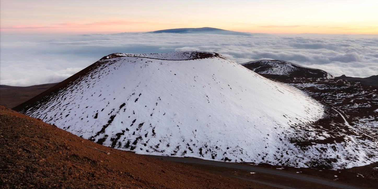 Snow-covered volcanic crater with reddish-brown slopes, surrounded by clouds below and distant mountains in the background at sunrise or sunset—a breathtaking scene that showcases one of the unique travel facts about the United States: this country is home to a remarkable variety of volcanic landscapes waiting to be explored.