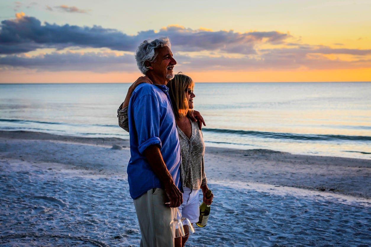An older couple stands on a sandy beach at sunset, smiling and embracing. The man holds empty wine glasses, and the woman carries a bottle, with the calm ocean and colorful sky in the background—enjoying a quiet moment away from the usual travel destinations boomers are skipping.