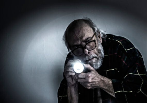 An older man with glasses holds a bright flashlight, wearing a plaid shirt. He leans forward and looks intently ahead, partially illuminated in a dark, shadowy environment.