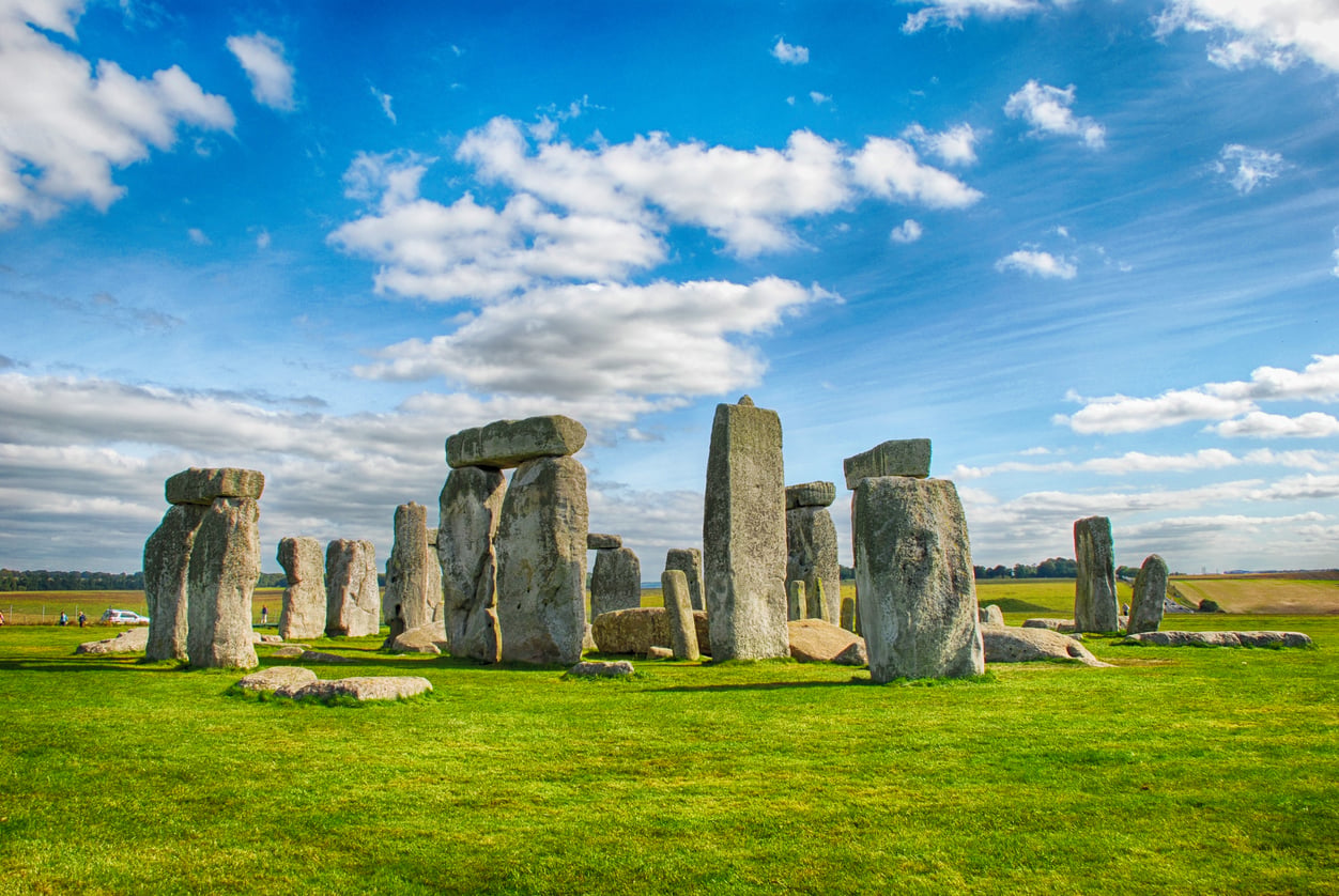 Ancient stone circle of Stonehenge under a bright blue sky with scattered clouds, surrounded by green grass and fields.