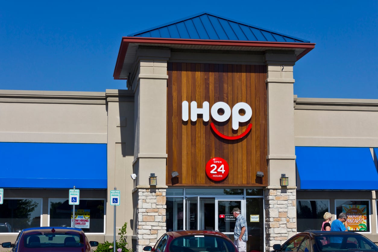 The front entrance of an IHOP restaurant with a large logo, blue awnings, and a sign indicating it is open 24 hours. Several parked cars and people are visible outside.