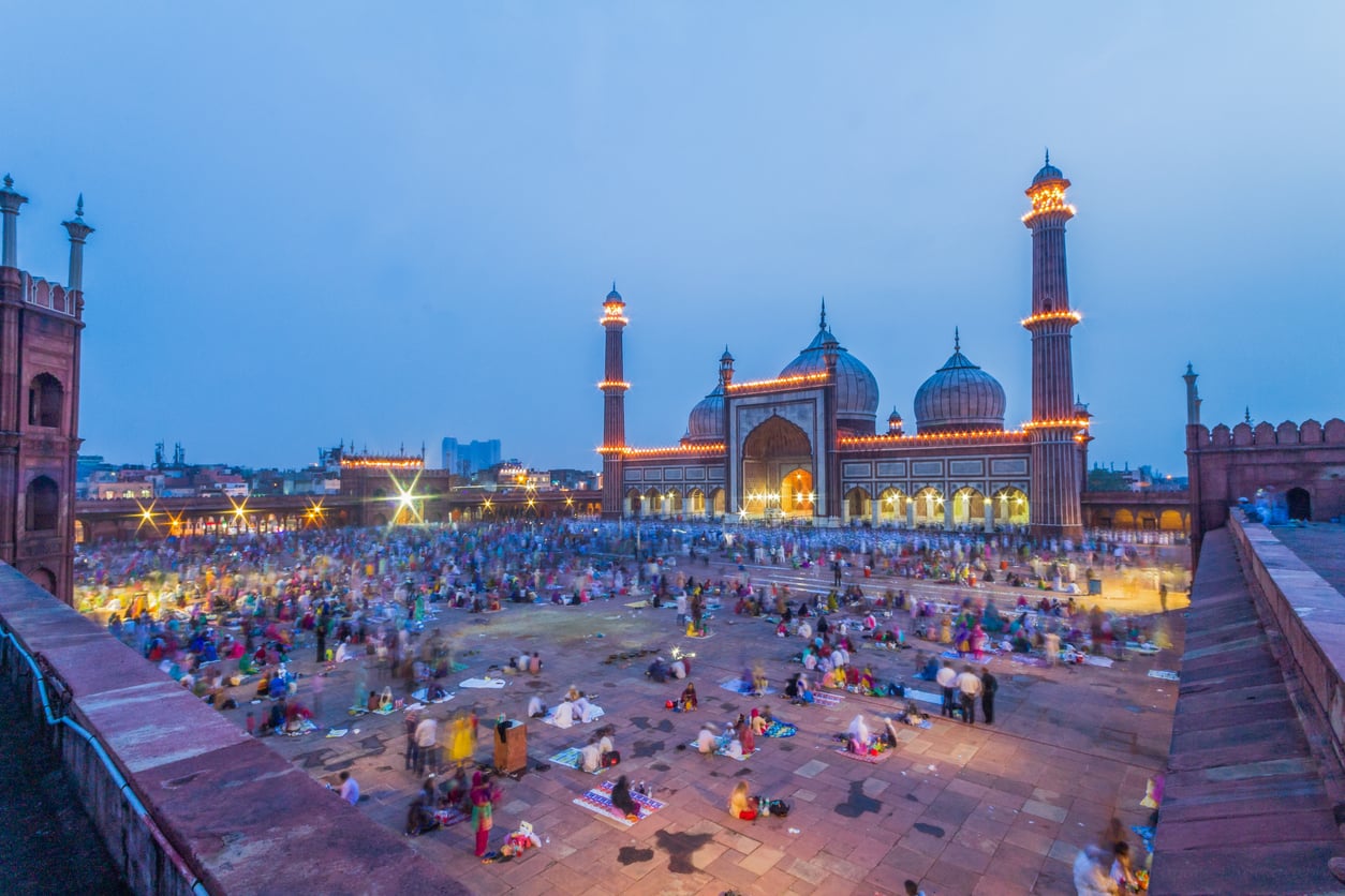 People gather at dusk outside a large, illuminated mosque with three domes and four minarets for what appears to be a communal event or prayer, possibly during Ramadan. Despite featuring on some lists of Places Travelers Are Warning Against Going In 2026, the sky is blue and the courtyard is bustling with activity.