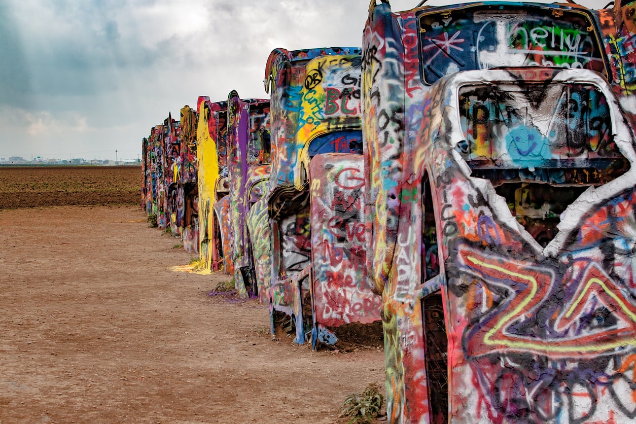 A row of half-buried, spray-painted cars stands in a dirt field under a cloudy sky, covered in colorful graffiti and vibrant designs.