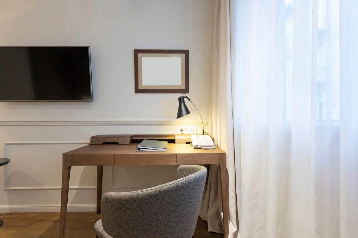 A modern hotel room desk with a lamp, phone, and open notebook—items reminiscent of things hotels used to offer guests—sits next to a light gray chair. A TV is mounted on the wall, while sheer curtains let in natural light from a window.