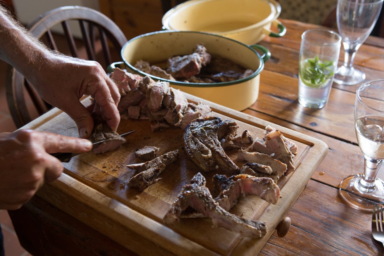 A person slices grilled meat on a wooden cutting board, with more cooked meat in a ceramic dish nearby. There are glasses of water and wine on a rustic wooden table.