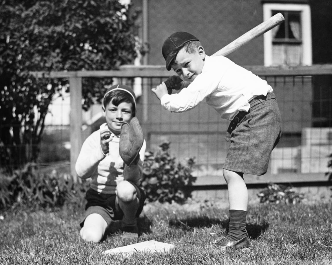 Two young boys play baseball in a yard. One boy kneels as the catcher with a glove, while the other stands ready to bat. Both wear shorts, long socks, and hats. There is a wooden fence and house in the background.