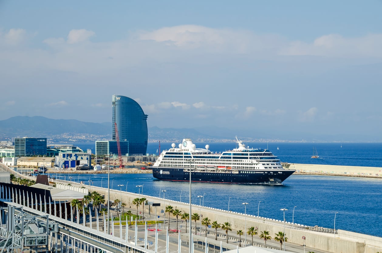 A large cruise ship docked at a harbor with the sea, palm trees, and modern buildings, including a tall glass hotel, in the background under a partly cloudy sky.