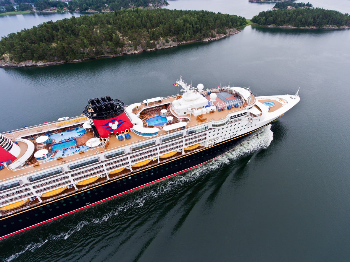 A large cruise ship with swimming pools on deck sails through calm water, surrounded by green, tree-covered islands under a cloudy sky.