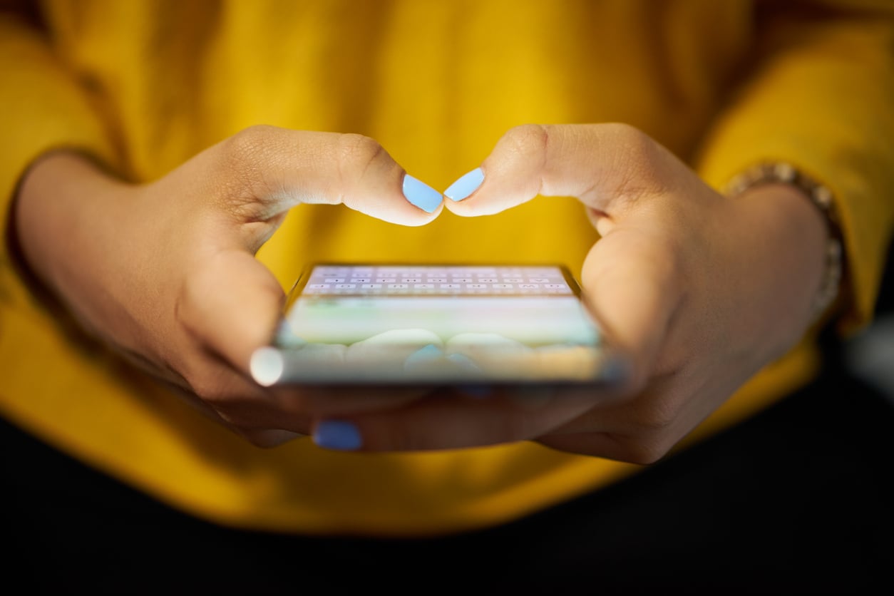 Close-up of hands with blue nail polish holding a smartphone, typing with thumbs. The person is wearing a yellow top and a beaded bracelet. The phone screen is illuminated.