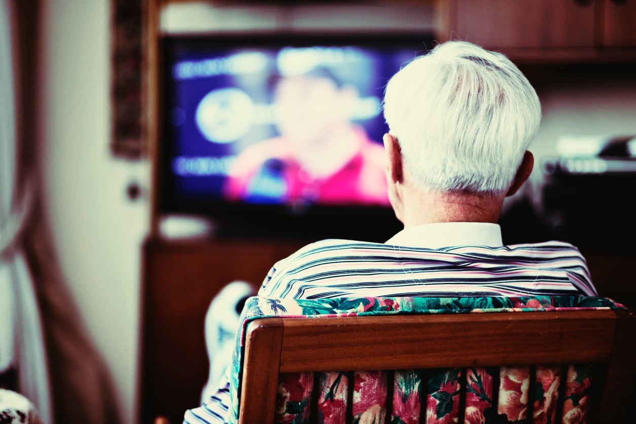 An elderly person with white hair sits on a floral chair, facing a blurry television screen in a cozy room. The focus is on the back of the person, showing a striped shirt and relaxed posture.