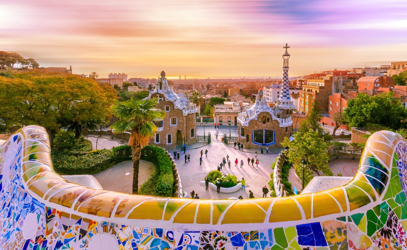 Colorful mosaic bench in Park Güell, Barcelona, overlooks whimsical Gaudà buildings and the city skyline at sunset, with people walking and bright, vibrant skies above—an iconic scene that stands in contrast to many travel destinations boomers are skipping these days.