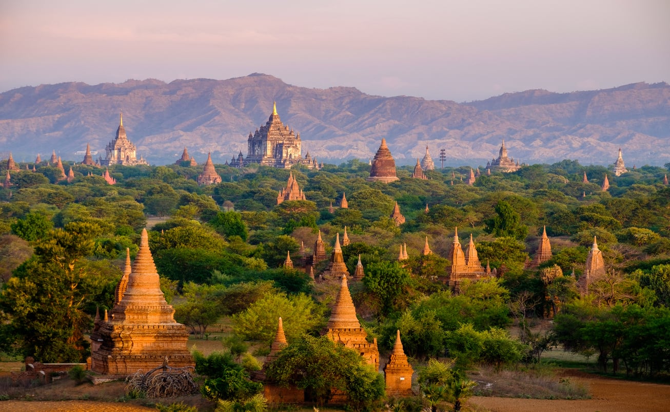 Ancient temples and pagodas rise amid lush greenery in Bagan, Myanmar, with distant mountains and a soft, golden evening light illuminating the landscape—a breathtaking scene that remains captivating even as Bagan appears on some lists of places travelers are warning against going in 2026.