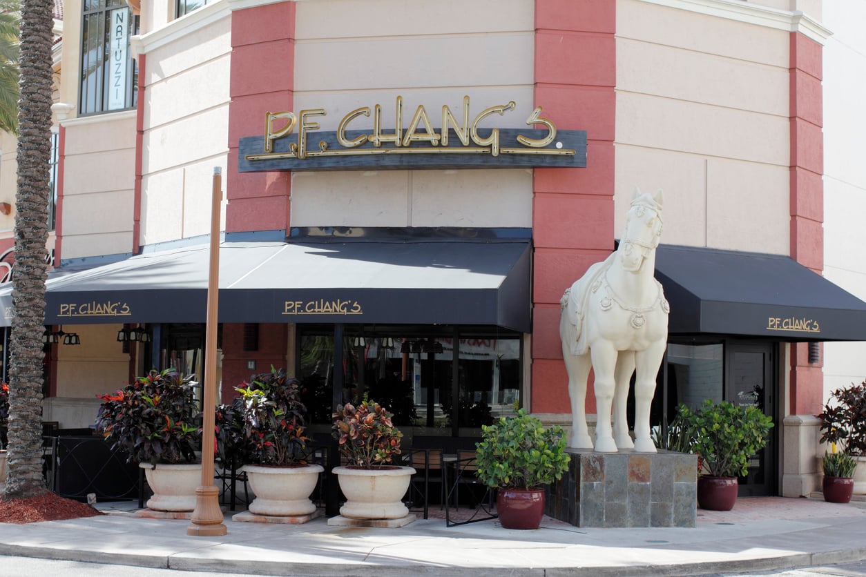 The exterior of a P.F. Chang’s restaurant featuring large windows, black awnings, potted plants, and a prominent white horse statue by the entrance.