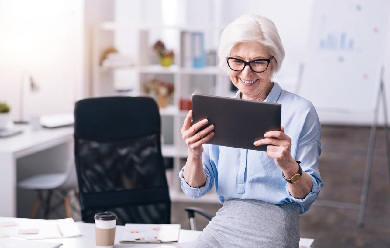 Smiling older woman with glasses sits at a desk in a modern office, holding and looking at a tablet. Reflecting Baby Boomer changes in the workplace, papers, a pen, and a takeaway coffee are on the desk nearby.