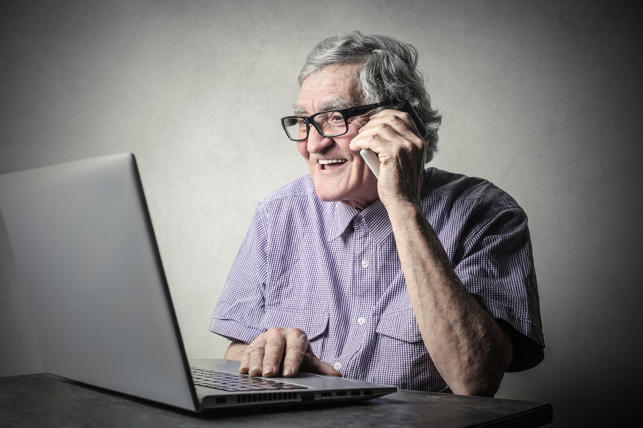 An older man wearing glasses and a purple shirt smiles while talking on a smartphone and using a laptop at a table, with a plain gray background.
