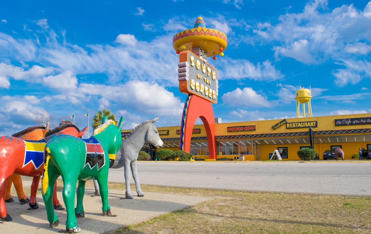 Colorful donkey statues stand in front of the large, sombrero-topped South of the Border sign and restaurant along a roadside under a bright blue sky with scattered clouds.