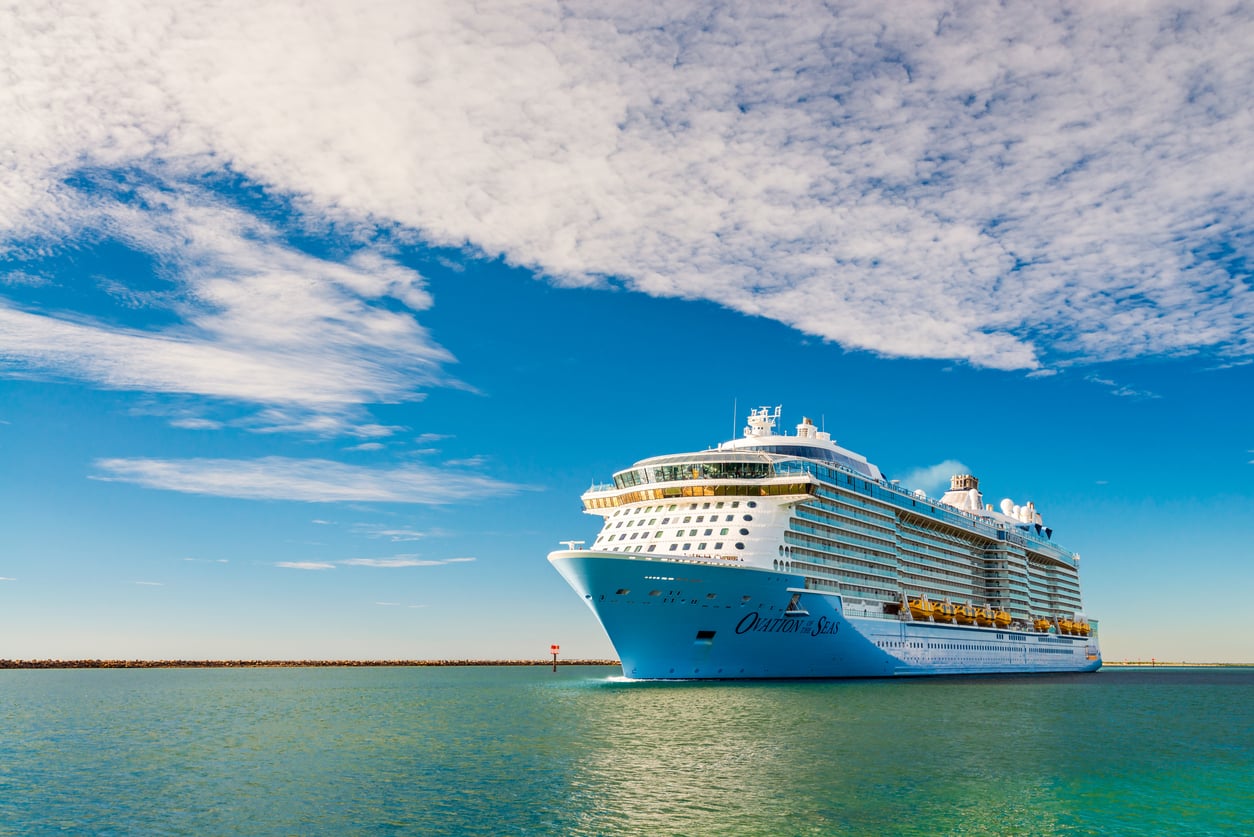 A large white cruise ship from one of the world’s premier cruise lines sails on calm blue-green water under a mostly clear sky with scattered clouds.