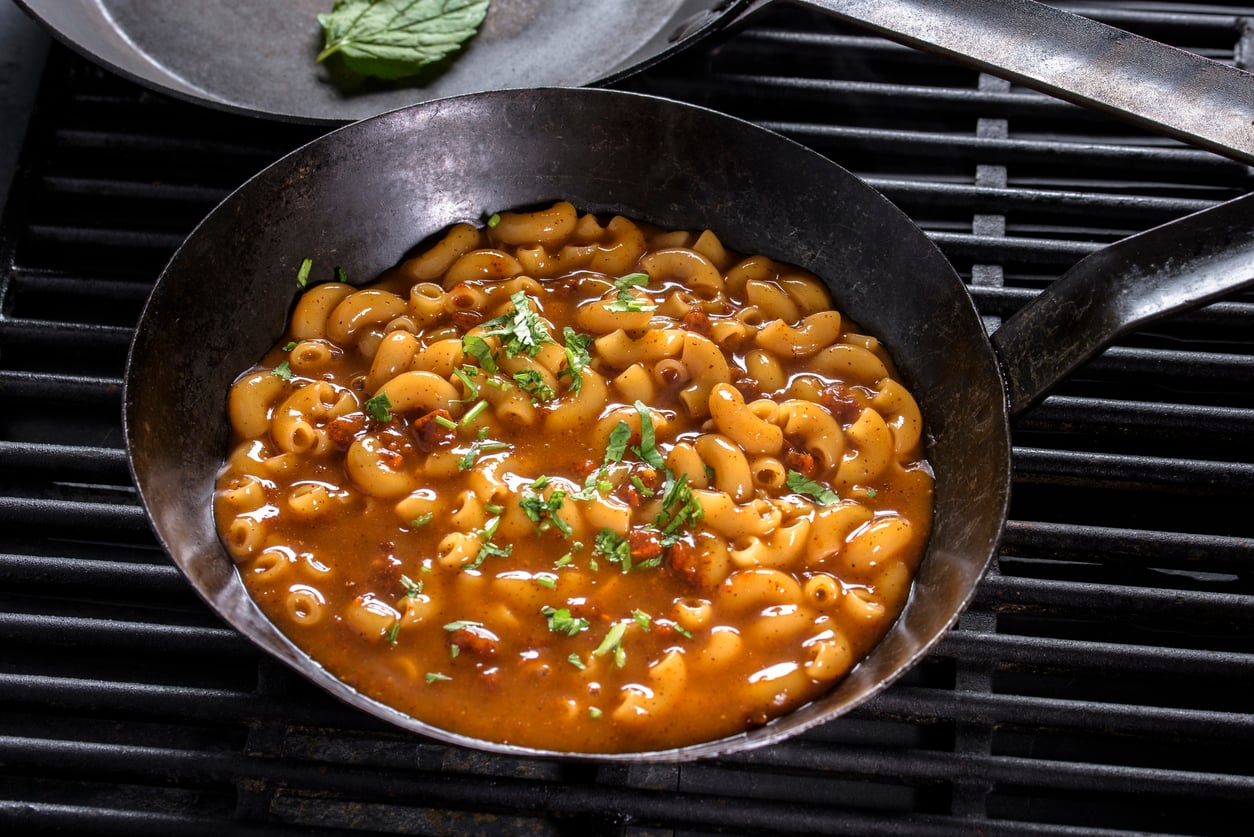 A pan with macaroni pasta in a rich, tomato-based sauce, reminiscent of the classic meals middle-class moms made, sits on a grill and is garnished with chopped herbs.