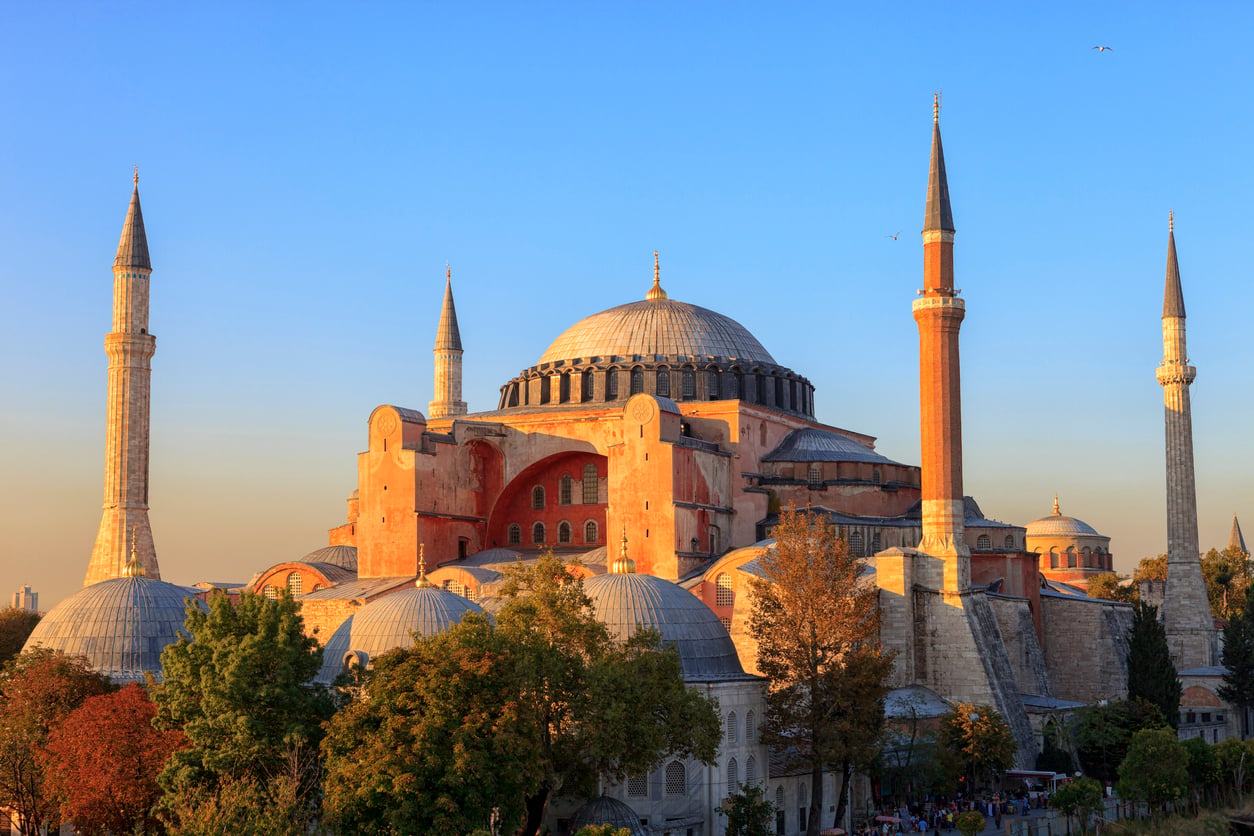 The Hagia Sophia in Istanbul, Turkey, is shown at sunset with its large central dome, four minarets, and surrounding trees under a clear blue sky.