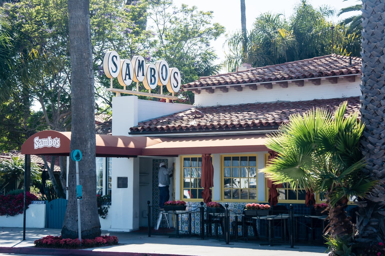 A restaurant with a tiled roof, yellow-framed windows, and a large “Sambos” sign on top. Outdoor seating is visible, with palm trees and greenery surrounding the building.
