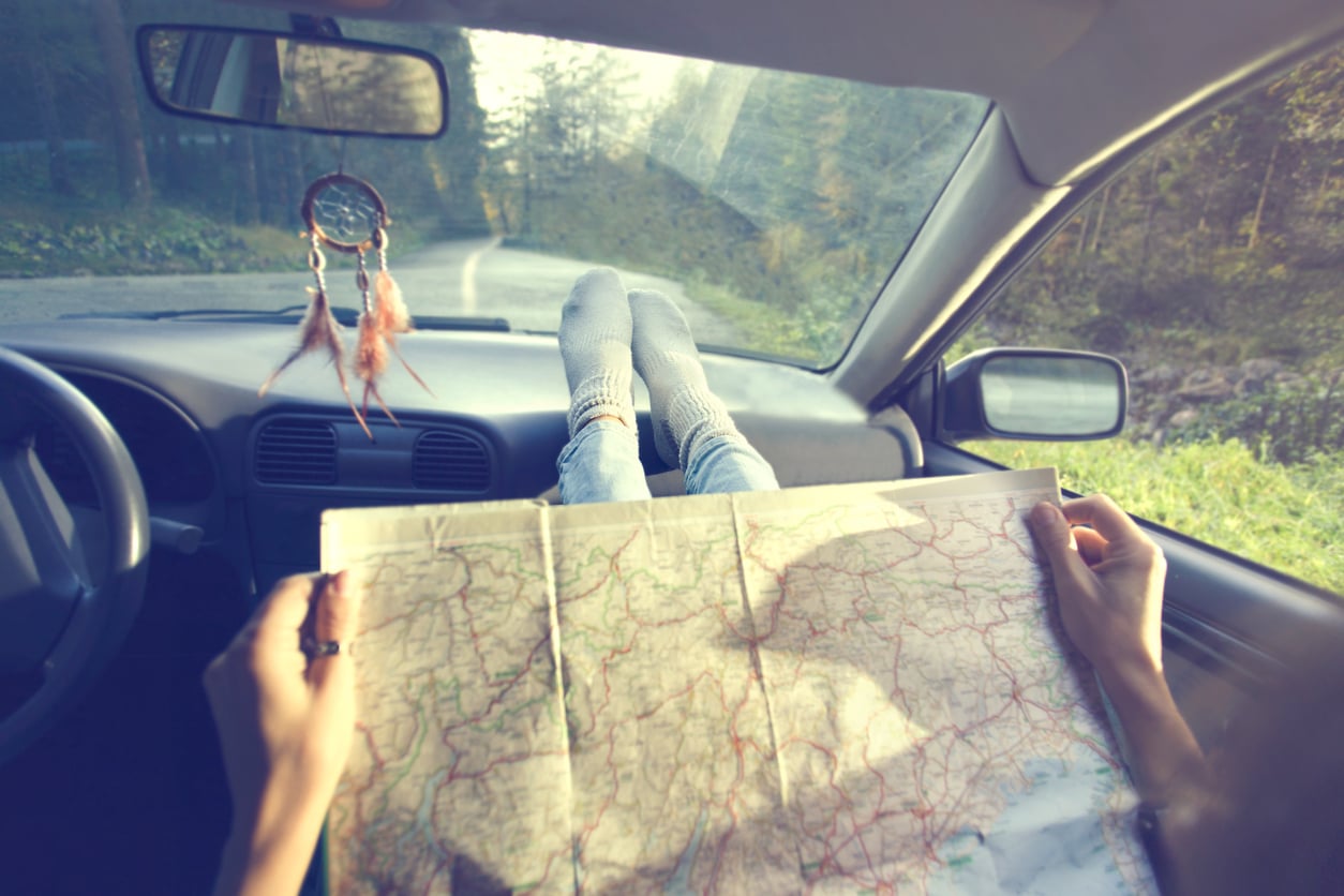 A person with socked feet on a car dashboard holds a large map. Sunlight streams through the windshield, and a dreamcatcher hangs from the rearview mirror. The car is parked on a tree-lined road.