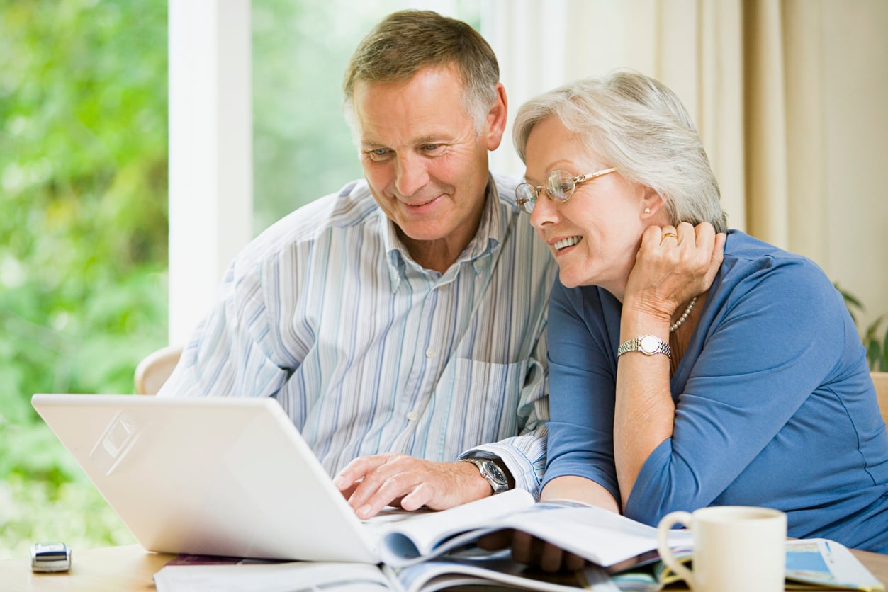 An older couple sits together at a table, smiling while looking at a laptop as they discuss their travel habits. Books and a mug are on the table, with a bright window and curtains in the background.