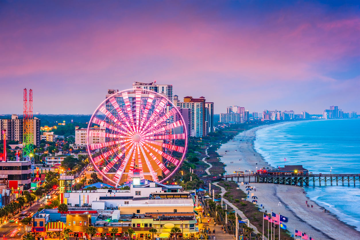 A brightly lit Ferris wheel glows near the beach at sunset, with city buildings and a vibrant skyline on one side and waves rolling onto the sandy shore on the other—a scene that perfectly captures why this destination is among the travel spots boomers are learning to love.