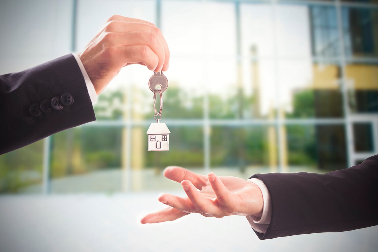 A person hands over a house-shaped keychain with keys to another person’s open hand, with a glass building in the background.
