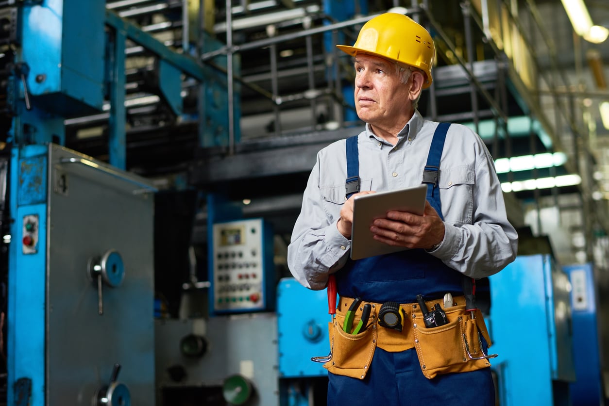 Older male worker in a yellow hard hat and tool belt holds a tablet, standing in an industrial factory with machinery and control panels in the background, perhaps reviewing retirement tips to plan for his next phase beyond the workplace.