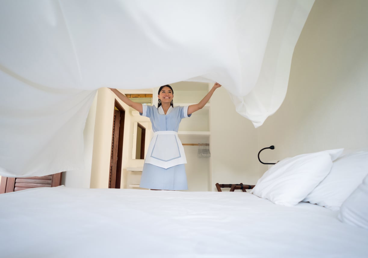 A person in a uniform smiles while lifting a white sheet to make a bed in a bright, tidy room with white walls and pillows—reminiscent of the exceptional attention to detail and warm service among the things hotels used to offer guests.