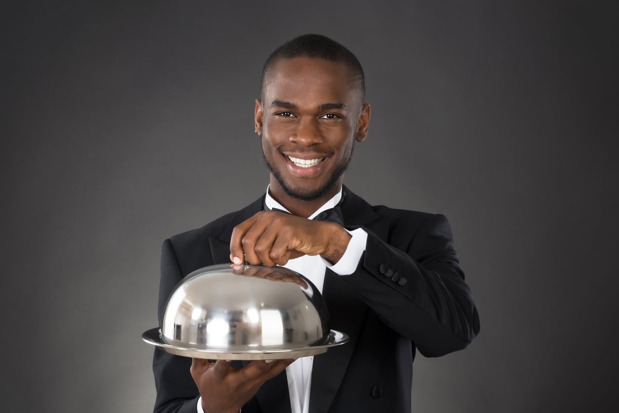 A smiling man in a tuxedo holds a silver serving tray with a domed lid against a dark background, as if presenting food.