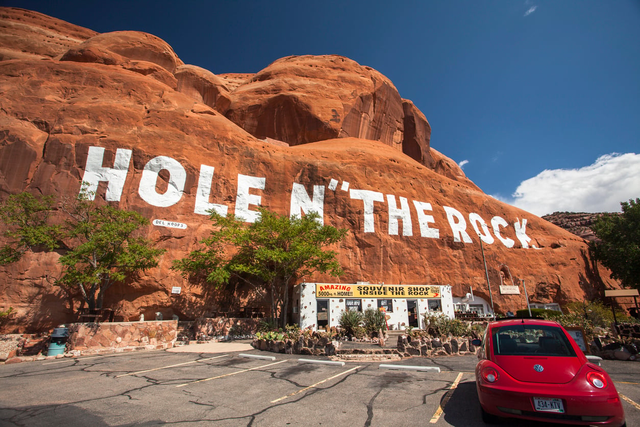 A red car parked outside the Hole N“ The Rock, a souvenir shop built into a large red sandstone rock formation under a bright blue sky.