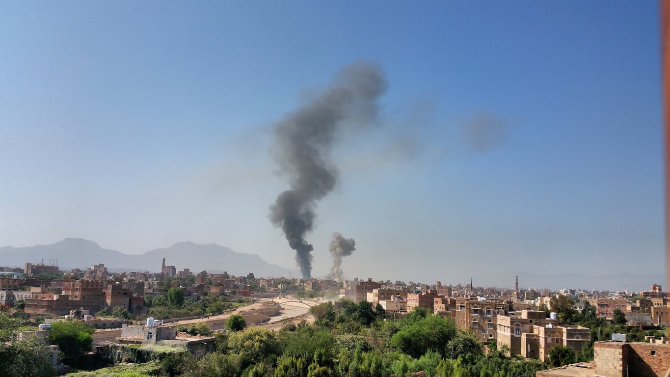 Thick black smoke rises from buildings in the distance of a city, with mountains in the background and clear blue sky above—a stark reminder of why this location has appeared on lists of places travelers are warning against going in 2026.