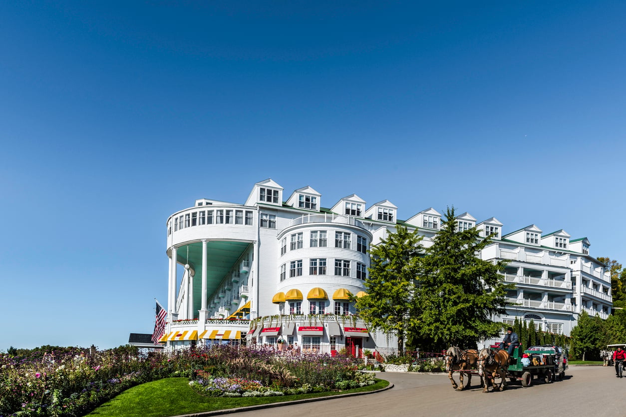 A large, elegant white hotel with dormer windows, listed among the most renowned Historic Hotels, sits surrounded by manicured gardens. Horse-drawn carriages pass by on a sunny day under a clear blue sky.