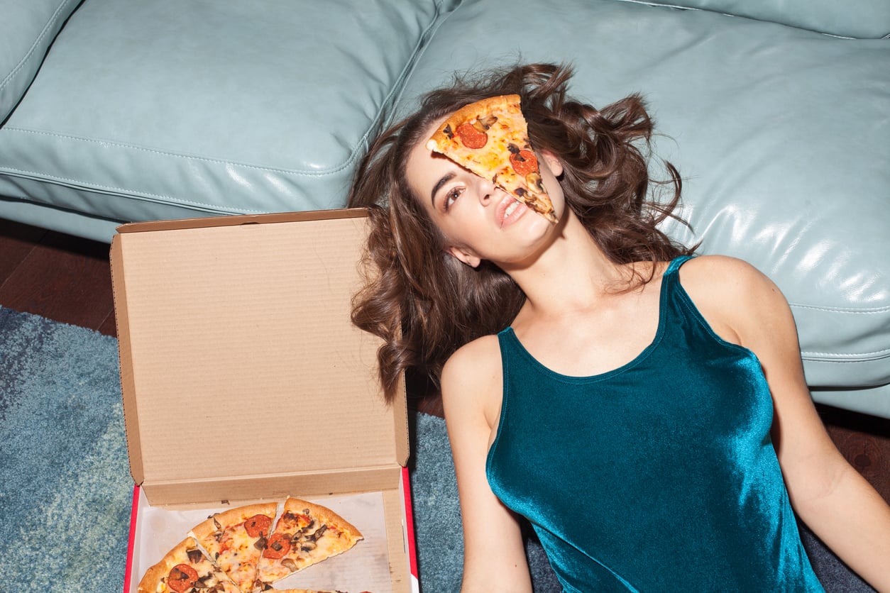 A woman in a teal dress lies on the floor next to a pizza box, with a slice of pizza resting on her face near her eyes and mouth.
