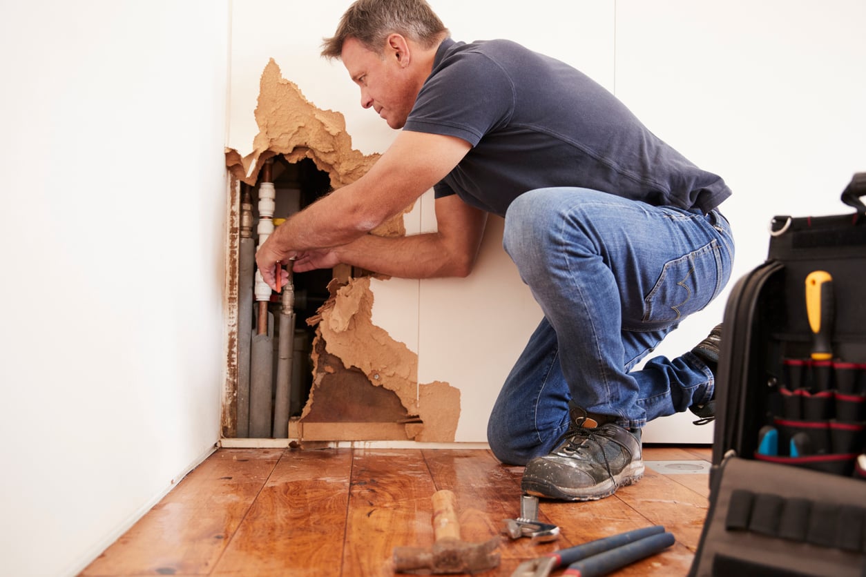 A man kneels on a wooden floor, repairing plumbing in a wall with damaged drywall. Tools and a tool bag are nearby.