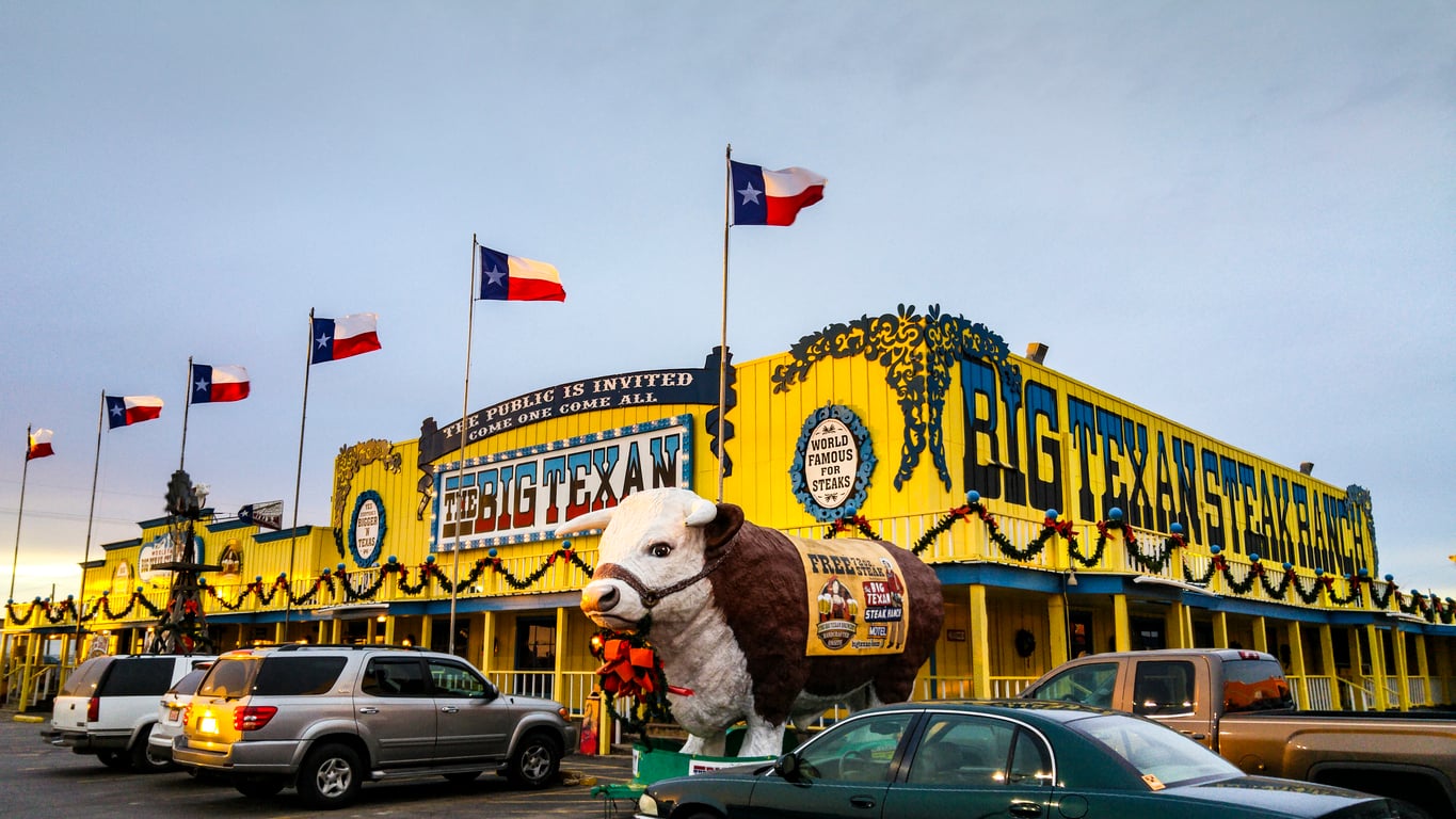A bright yellow steakhouse with Texas flags, “Big Texan” signs, and a large cow statue in front embodies the spirit of Classic Roadside Attractions. Cars are parked in the lot, and garlands decorate the building.