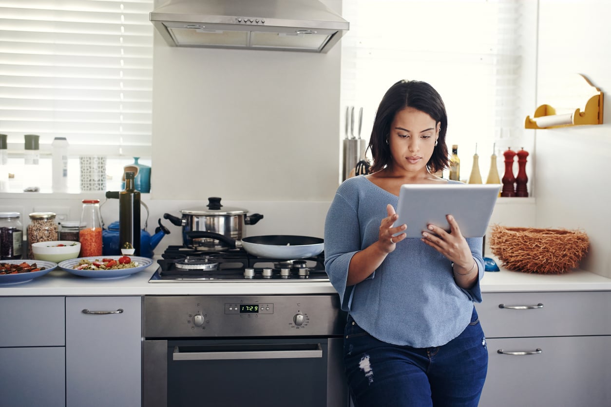 A woman stands in a modern kitchen, leaning against the counter while looking at a tablet. Food and kitchen utensils are on the counter beside her, and sunlight streams in through the window.