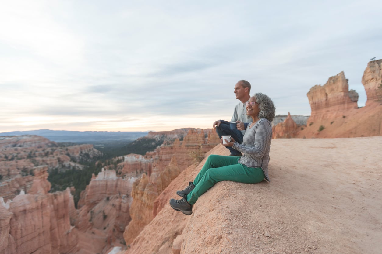 Two people sit at the edge of a rocky cliff, enjoying the scenic view of a canyon with layered rock formations and a cloudy sky in the background.