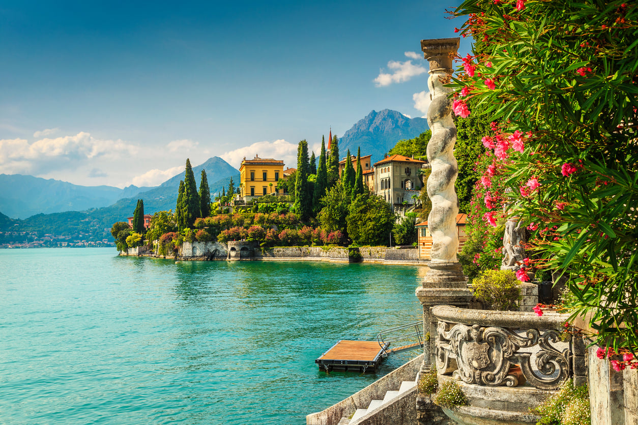 A scenic lakeside view of colorful villas, lush green trees, and flowering plants with mountains in the background and turquoise water in the foreground, likely depicting Lake Como, Italy—a stunning spot among the travel destinations boomers are skipping.