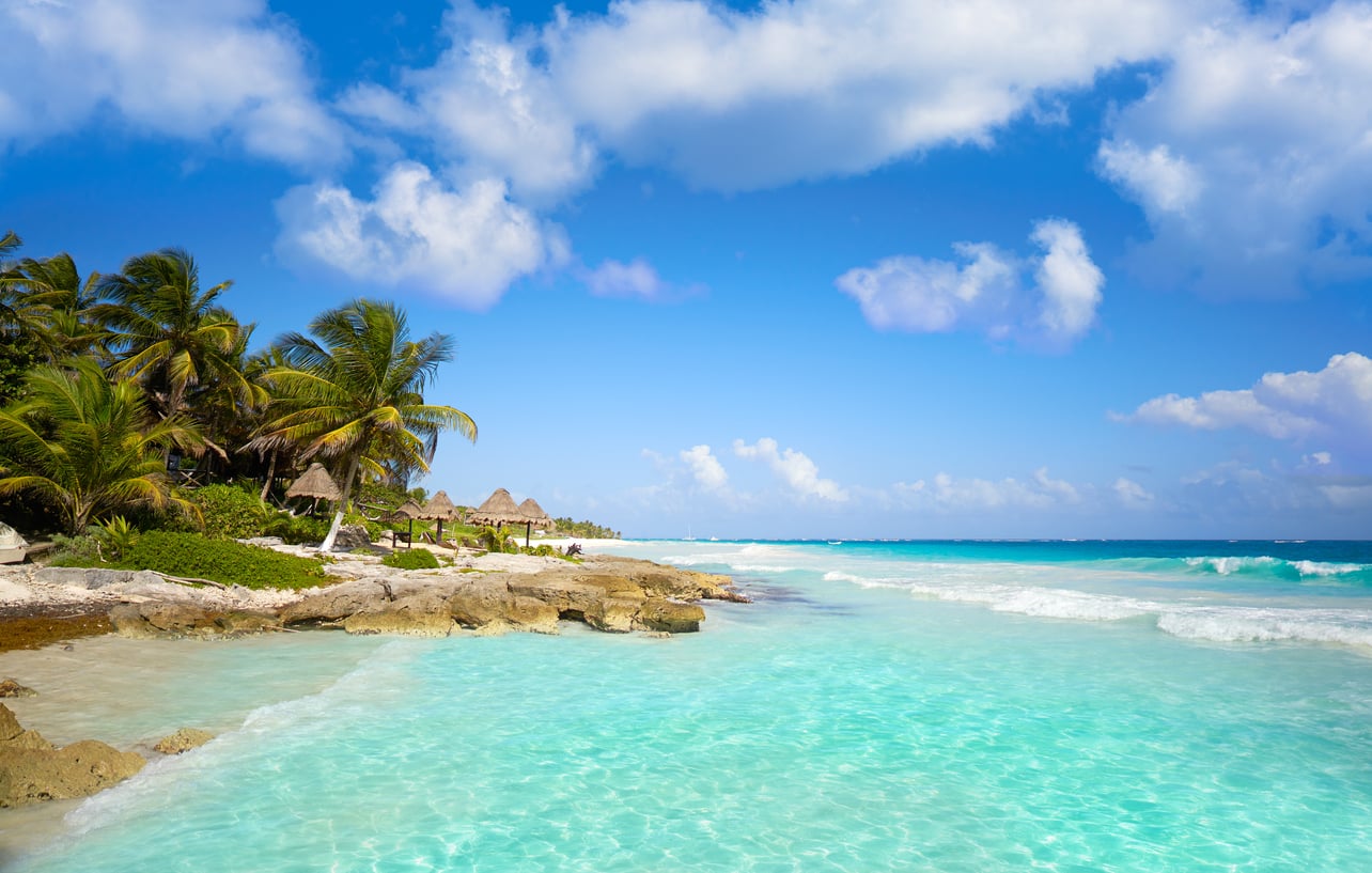Tropical beach with clear turquoise water, palm trees, rocky shoreline, and thatched huts under a blue sky with scattered clouds—one of the stunning travel destinations boomers are skipping in favor of quieter escapes.