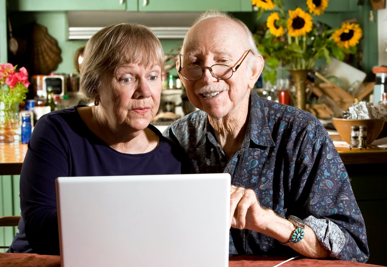 An elderly couple sits closely together at a table, looking at a laptop screen. They appear focused and engaged, perhaps exploring retirement tips online. The background shows a kitchen with sunflowers and colorful decor.