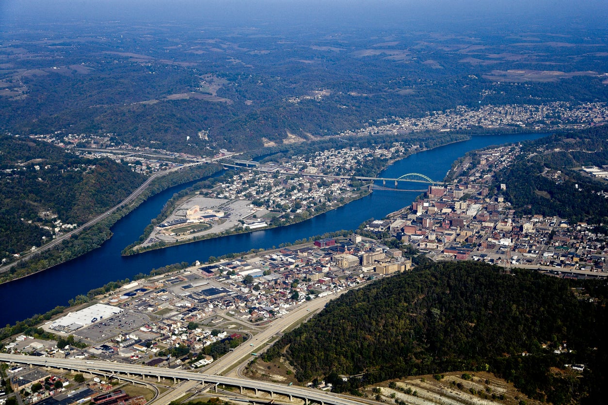 Aerial view of a river winding through a city with bridges, buildings, roads, and green hills in the background under a clear sky.