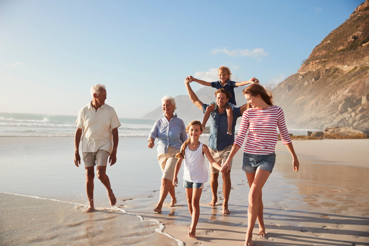 Three adults, two children, and two seniors—each with their own unique travel habits—walk and smile together along a sandy beach by the ocean, with cliffs in the background on a sunny day.