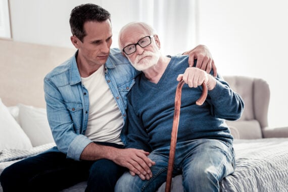 A younger man sits beside an older man with a cane on a bed, offering comfort with his arm around him. Both look thoughtful and serious in a softly lit room, reflecting on the Baby Boomer changes that have shaped their lives and relationship.